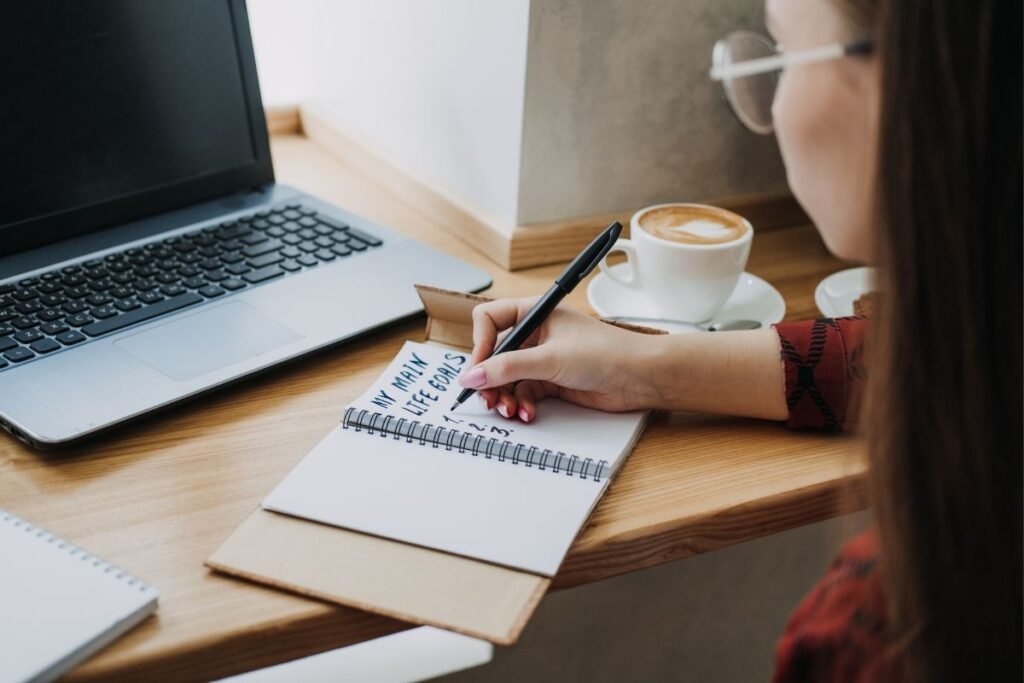 Uma mulher escrevendo "My Main Life Goals" em um caderno aberto sobre uma mesa com um laptop e uma xícara de café.