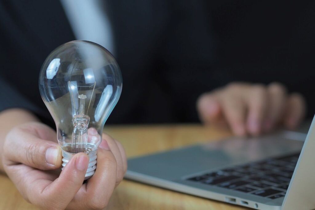 Uma mão segura uma lâmpada acesa, simbolizando uma ideia ou inovação, com um laptop desfocado ao fundo, representando a conexão entre criatividade e tecnologia no empreendedorismo.