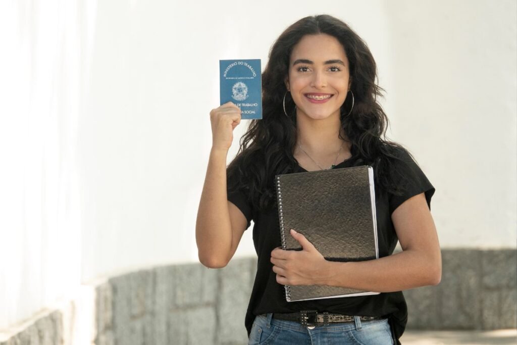 Jovem feliz segurando uma Carteira de Trabalho brasileira e um caderno, representando a conquista do primeiro emprego.