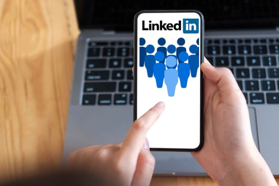 Mãos segurando um smartphone com o aplicativo do LinkedIn aberto, exibindo ícones de pessoas em azul sobre um fundo branco, com um teclado de laptop em segundo plano.