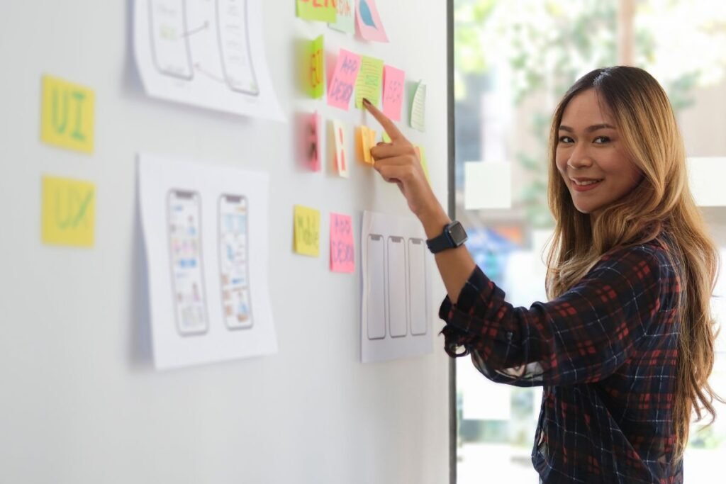 Mulher sorridente interagindo com um quadro branco cheio de notas adesivas e esboços de interfaces de aplicativos, representando o planejamento de um projeto.