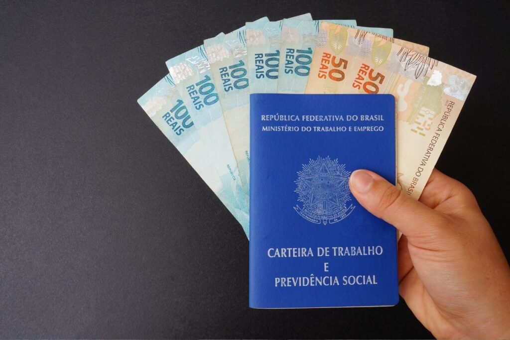 Mão segurando Carteira de Trabalho e Previdência Social com notas de cem e cinquenta reais, representando o 13º salário.