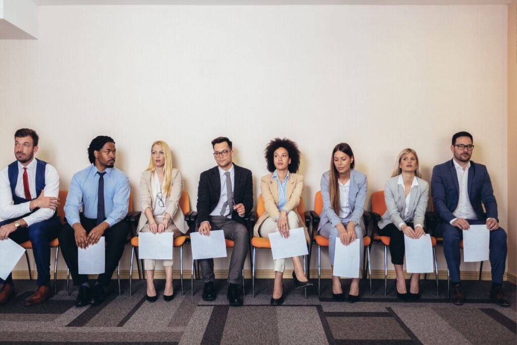Grupo diverso de candidatos aguardando em cadeiras uma entrevista de emprego, segurando currículos.
