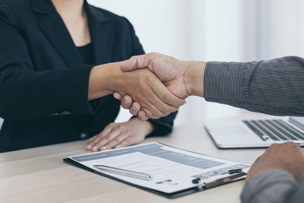 Aperto de mãos cordial entre recrutador e candidato após uma entrevista de emprego, com currículo na mesa.