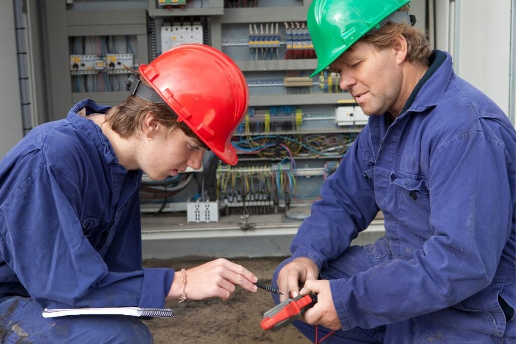Dois eletricistas, um jovem aprendiz e um profissional, trabalhando com equipamentos elétricos.