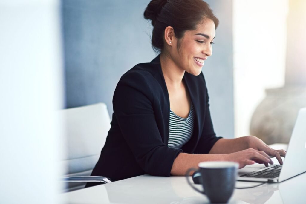 Mulher sorridente trabalhando no laptop com uma xícara de café ao lado.