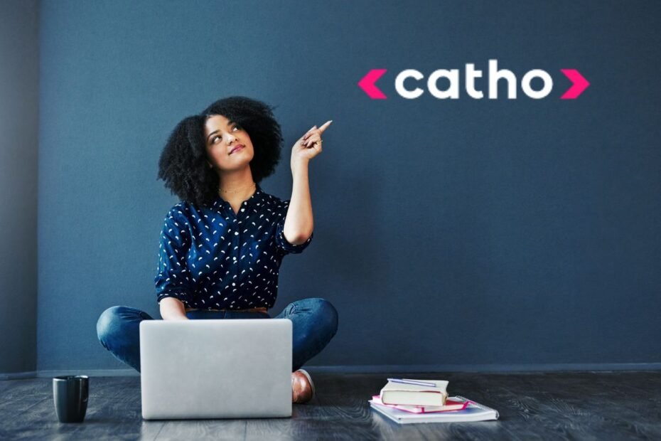 Jovem profissional sorridente apontando para logo da Catho enquanto usa laptop para criar currículo