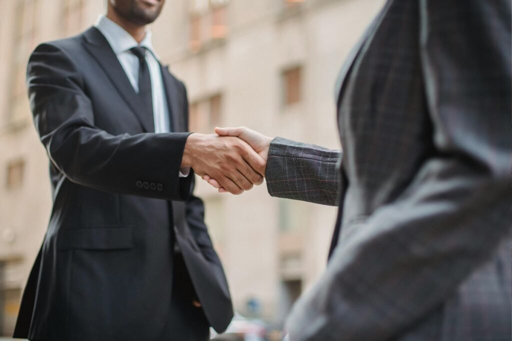 Duas pessoas de negócios, vestindo ternos, apertando as mãos em um ambiente urbano, simbolizando uma nova parceria ou acordo.
