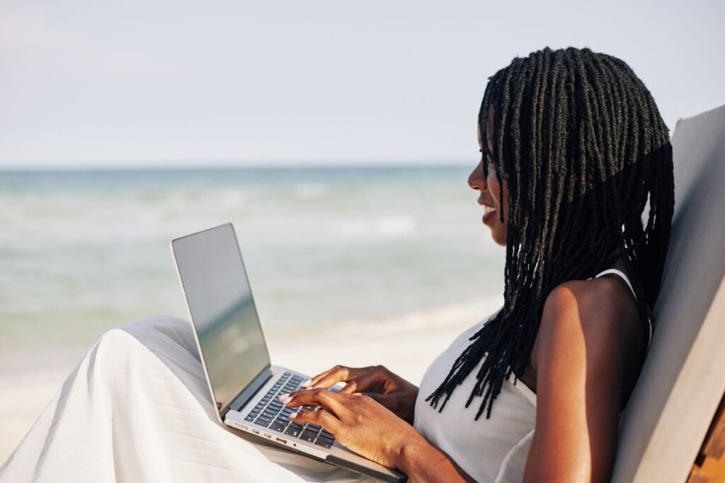 Mulher negra com dreadlocks usando um laptop em uma espreguiçadeira na praia, com o mar ao fundo, simbolizando o trabalho remoto ou a flexibilidade do descanso.