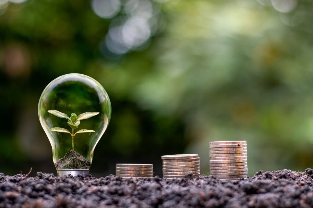 Uma lâmpada transparente com uma pequena planta brotando dentro, ao lado de pilhas crescentes de moedas sobre terra escura, com um fundo verde desfocado.