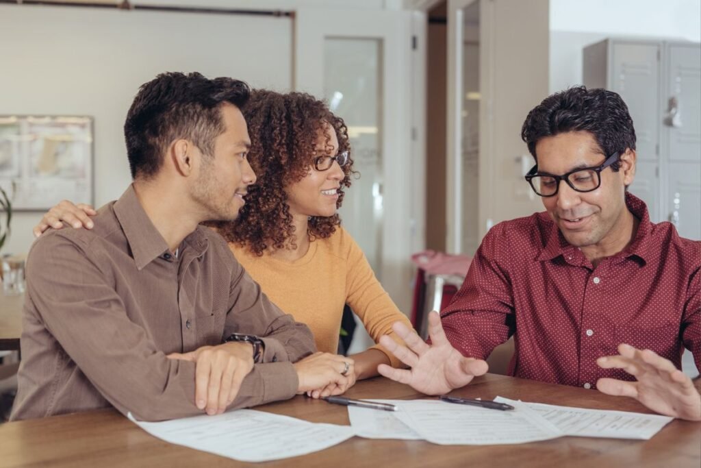 Um casal atento ouvindo um consultor financeiro ou mentor, que gesticula enquanto explica algo sobre documentos na mesa.