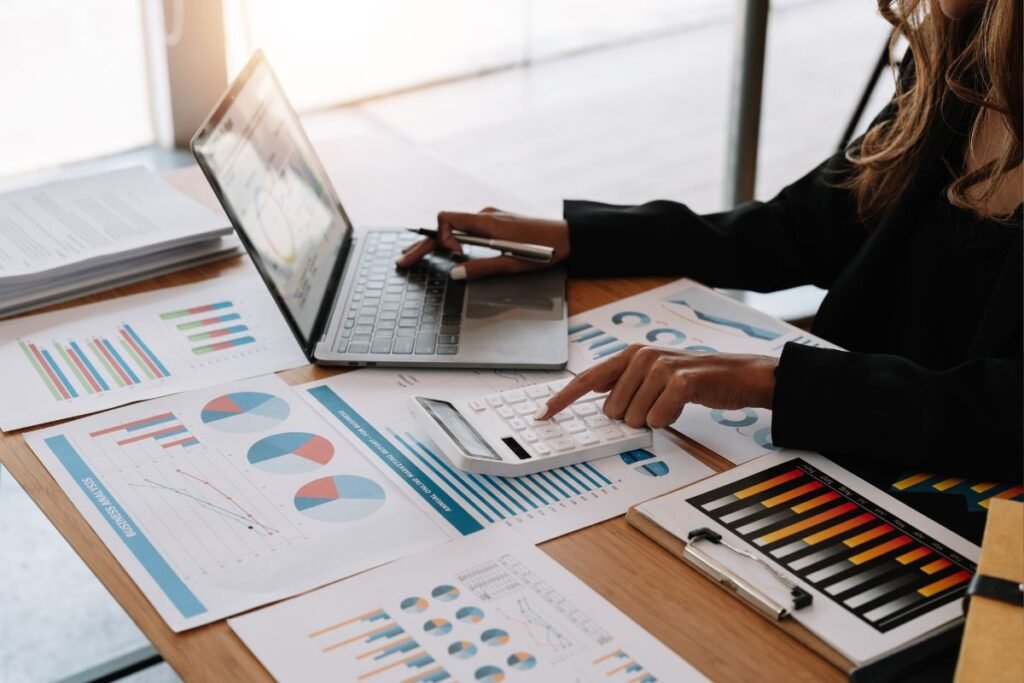 Mulher de negócios analisando dados financeiros e gráficos em um laptop e calculadora, cercada por relatórios de análise de negócios.