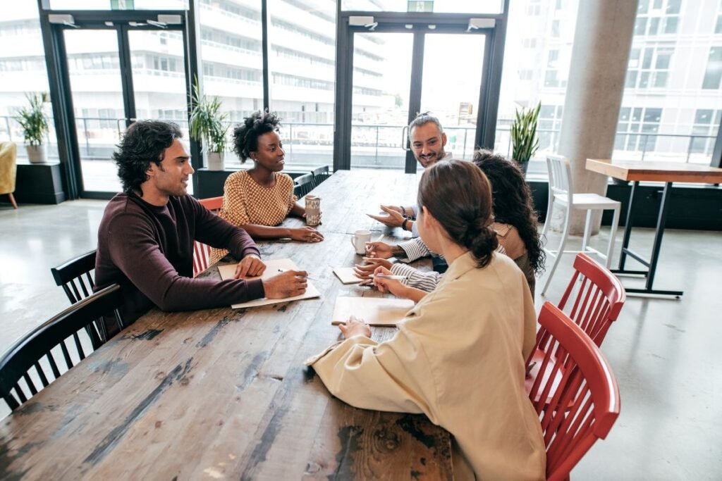 Um grupo diverso de cinco pessoas sentadas em torno de uma grande mesa de madeira, conversando e interagindo em um ambiente de escritório moderno com grandes janelas ao fundo.