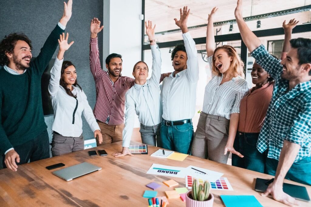 Um grupo diversificado de sete pessoas, homens e mulheres, com os braços erguidos em celebração, em torno de uma mesa de reunião com materiais de branding. Todos sorriem e parecem estar comemorando uma conquista.
