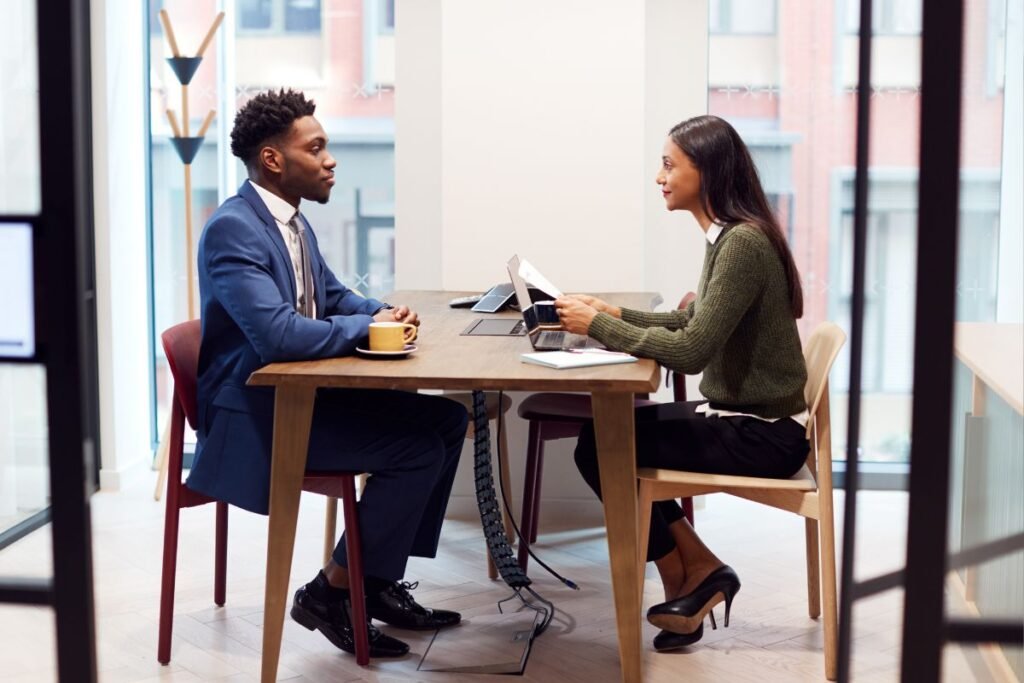 Um homem e uma mulher, ambos profissionais, sentados à mesa um de frente para o outro durante uma entrevista de emprego. O homem, em um terno azul, está sendo entrevistado pela mulher, vestida casualmente elegante, em um ambiente de escritório moderno com luz natural e janelas amplas.